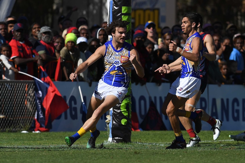 An AFL player runs away from goal in celebration after scoring, as his teammate watches him.