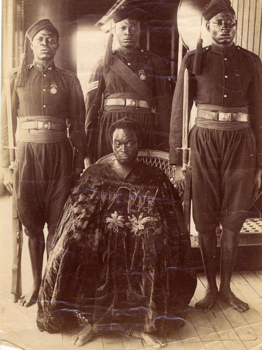 The Benin King sits in a chair wearing a velvet gown. His feet are shackled. Behind him are three soldiers with bayoneted rifles