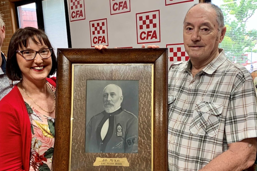 a married couple in their fifties stand with a couple in their eighties, holding a photo of their relative James McKay