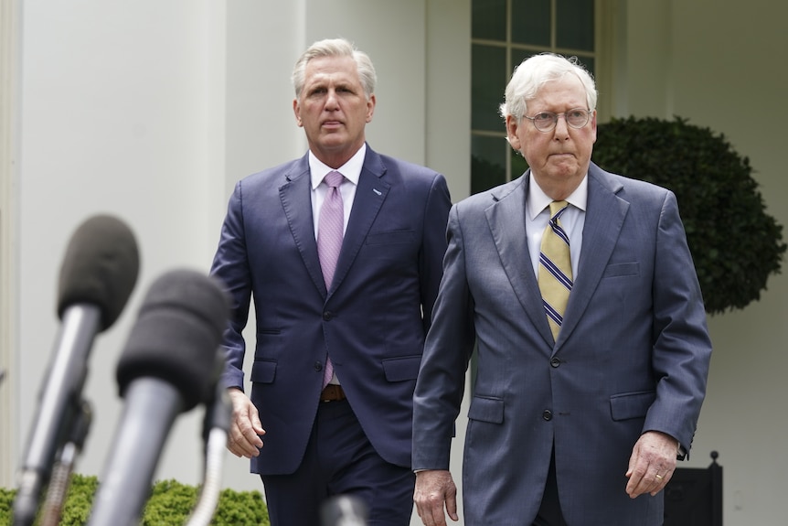 Mitch McConnell and Kevin McCarthy, both wearing suits, walk towards microphones