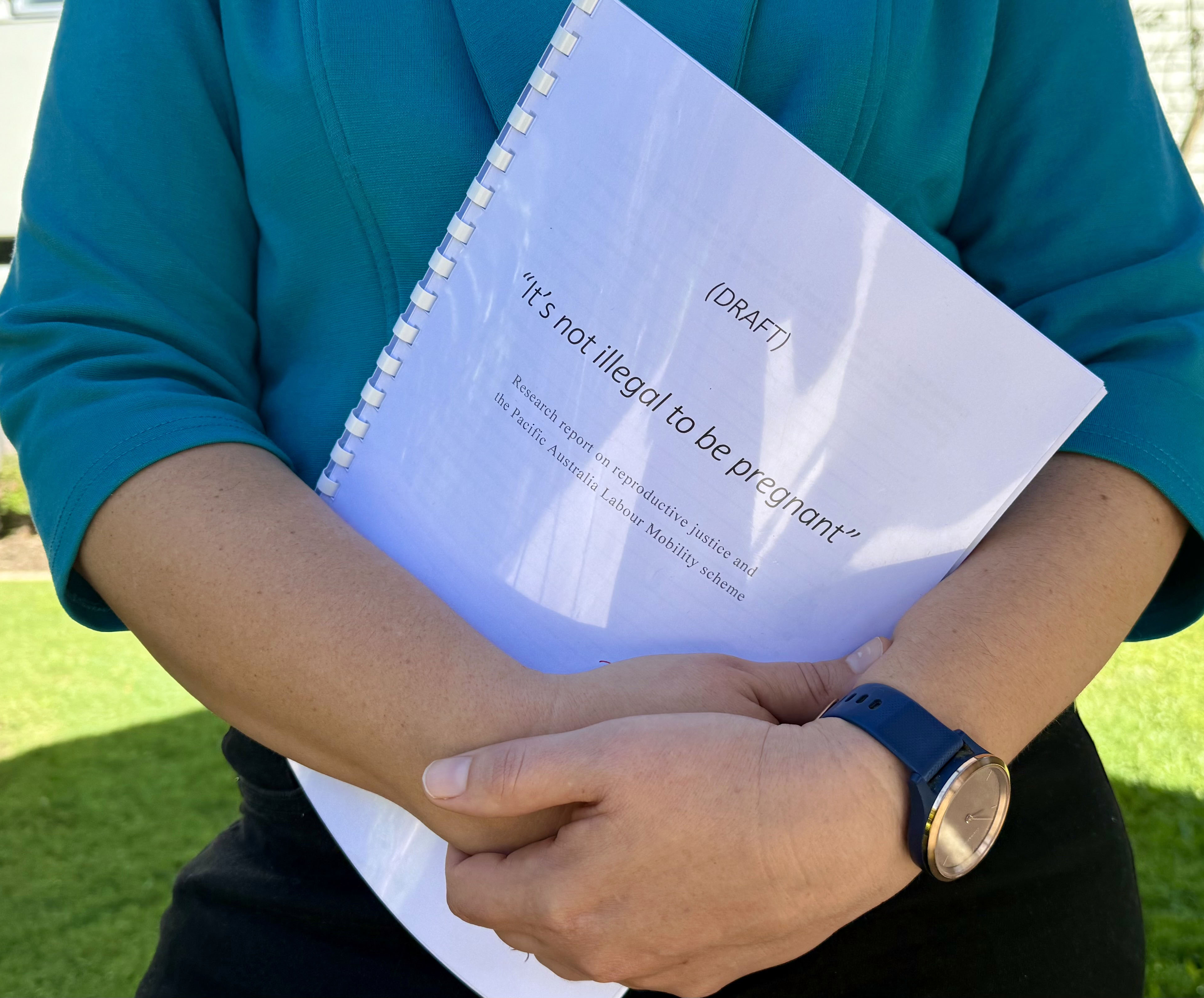 A woman's hands hold a bound report. The white paper title page says ' DRAFT - It's not illegal to be pregnant."