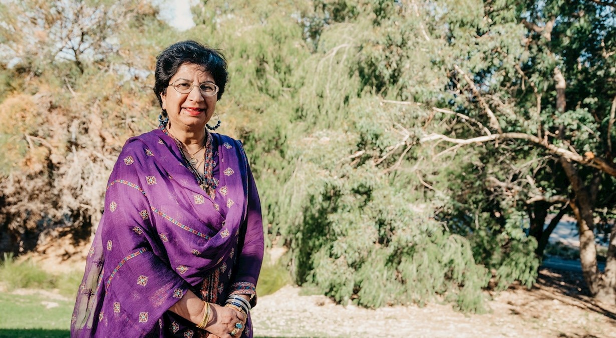 An older Pakistani woman in a patterned, purple sari. She has short, dark hair and glasses and is standing near bushland.