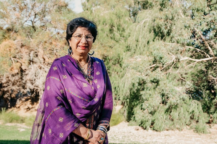 An older Pakistani woman in a patterned, purple sari. She has short, dark hair and glasses and is standing near bushland.