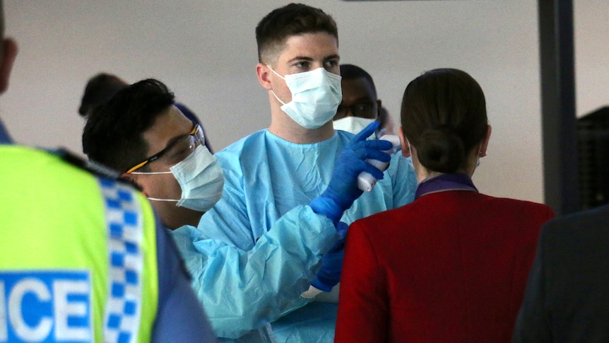 A flight attendant has her temperature checked at Perth Airport by two health workers wearing PPE, with a police officer nearby.