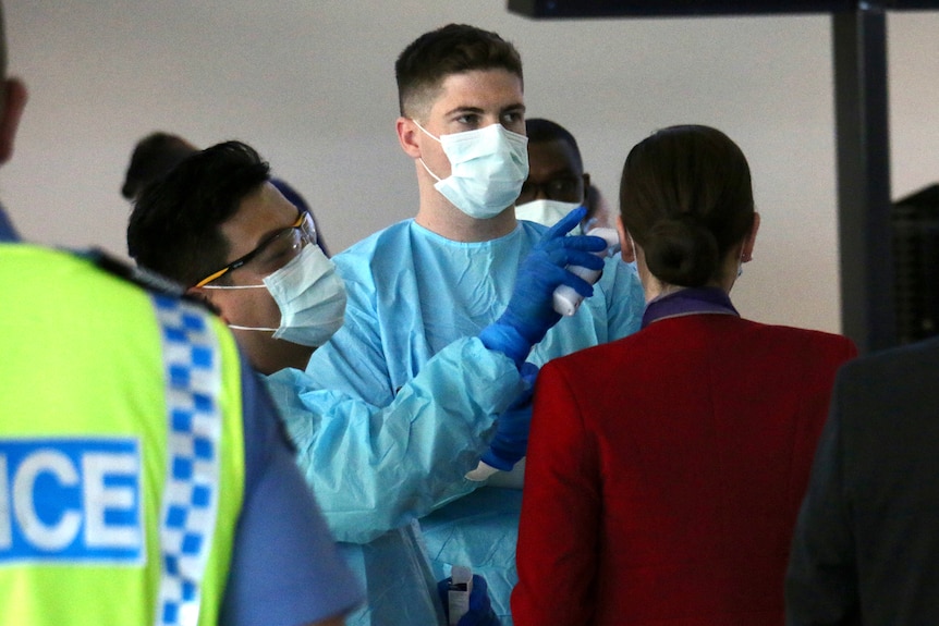 A flight attendant has her temperature checked at Perth Airport by two health workers wearing PPE, with a police officer nearby.