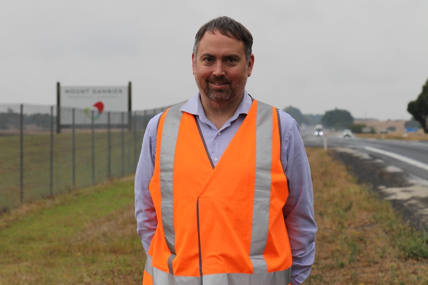 A man in a high-vis vest smiles at the camera with a sign, fenceline and highway stretching behind him.