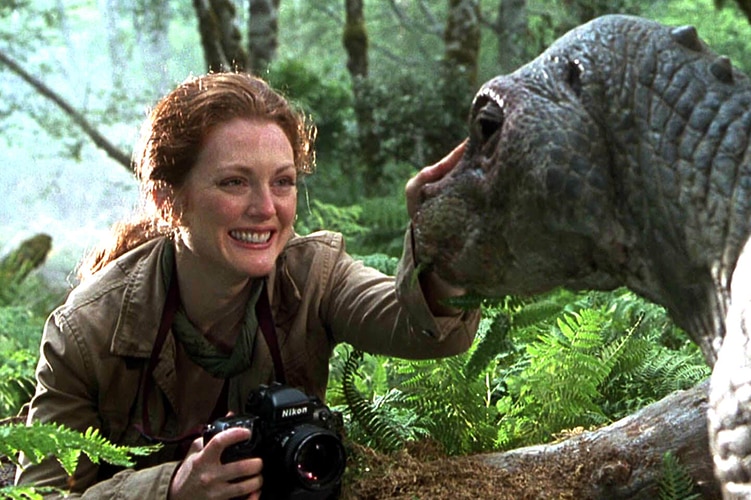A woman with long red hair holds camera, smiles and pats dinosaur like creature in jungle on bright day.
