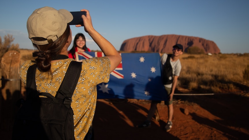 A girl takes a photo of two people with an Australian flag near Uluru. The girl's back is turned.