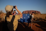 A girl takes a photo of two people with an Australian flag near Uluru. The girl's back is turned.