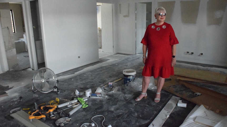 Woman in bright red dress stands in her demolished house and looks out to the pool