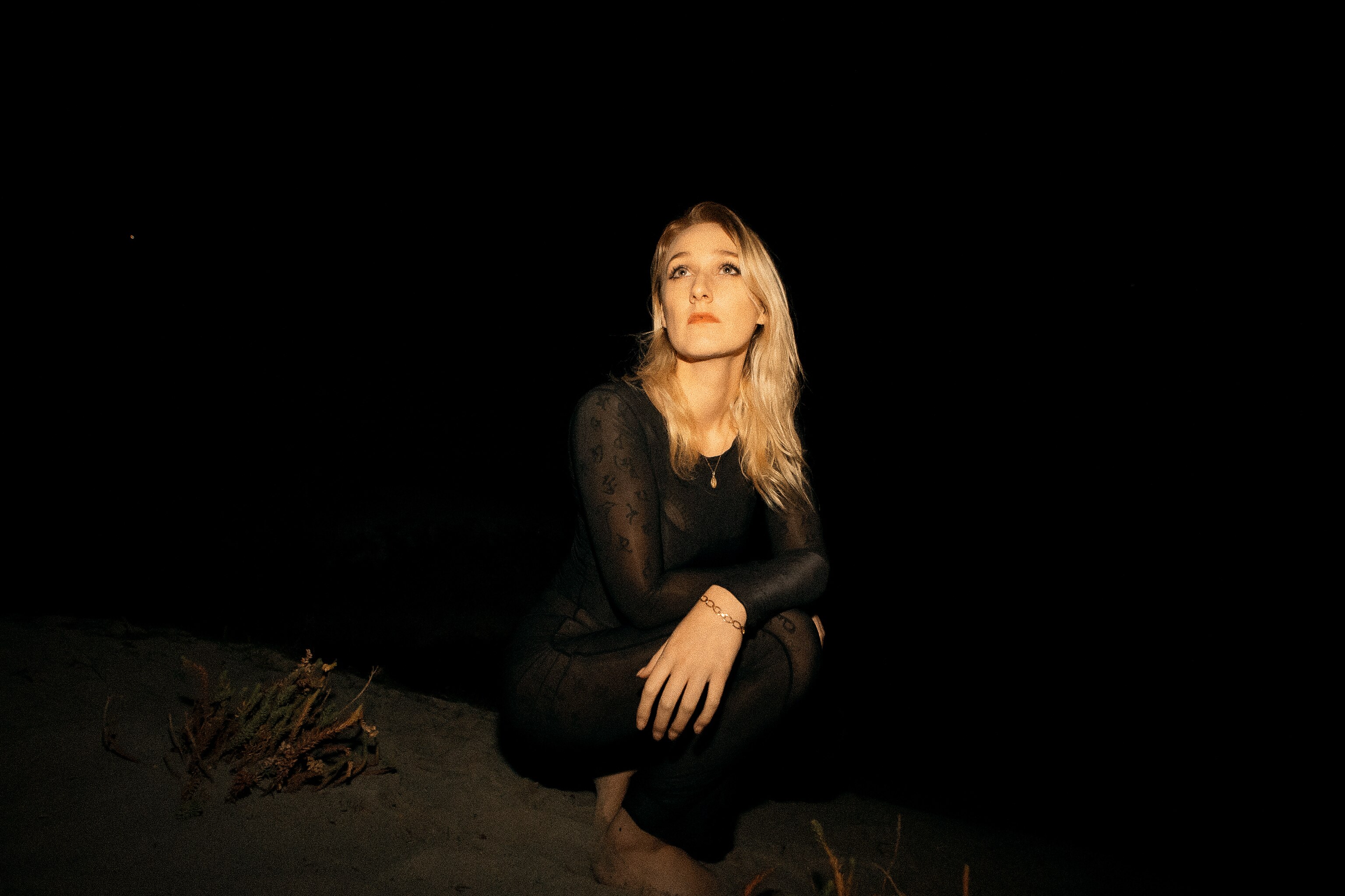 Carla Geneve crouches down on the ground and looks to the sky. She is outdoors and the background is dark.