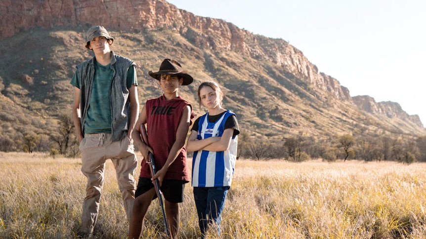 Three young people stand in front of a outback landscape, the youngest holding a rifle.