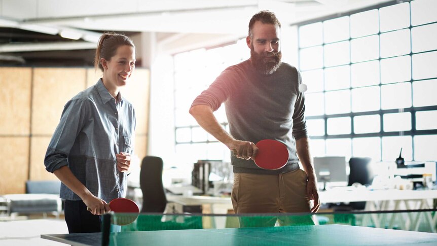 Within a large office space, a woman and man stand at one end of a tabletennis table, holding bats and smiling.