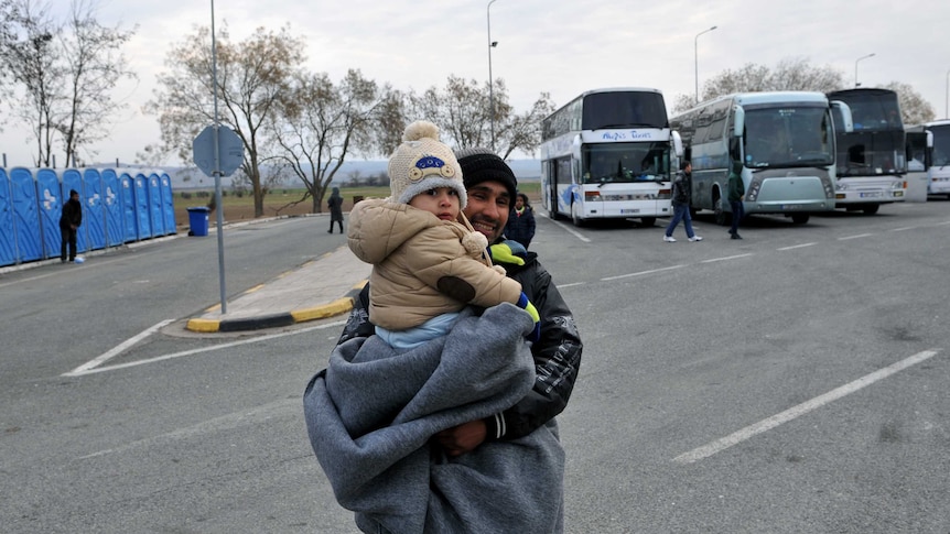 An asylum seeker carrying his child stand next to buses as he waits to cross the Greek-Macedonian border.