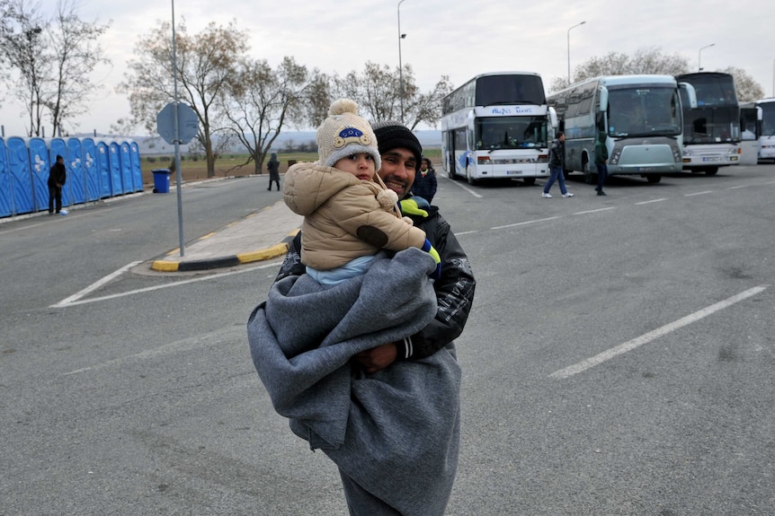 An asylum seeker carrying his child stand next to buses as he waits to cross the Greek-Macedonian border.