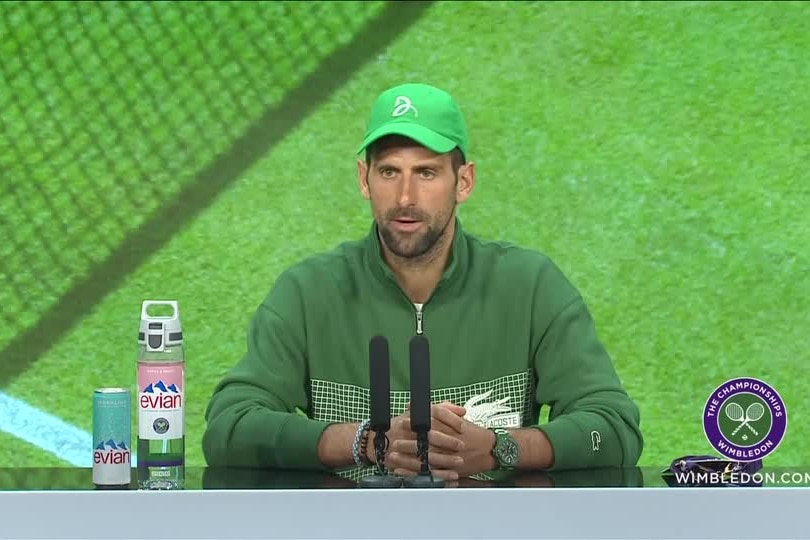 Novak Djokovic wearing a forest green sweatshirt, baseball cap and watch with a green tennis ground background