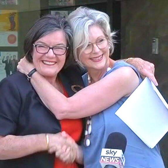 Two woman embrace and shake hands in front of a microphone at an outdoors media conference.