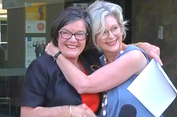 Two woman embrace and shake hands in front of a microphone at an outdoors media conference.