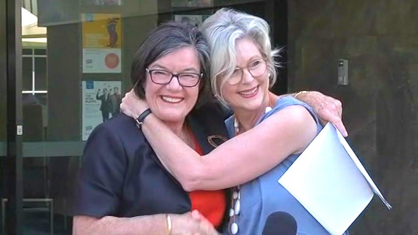 Two woman embrace and shake hands in front of a microphone at an outdoors media conference.