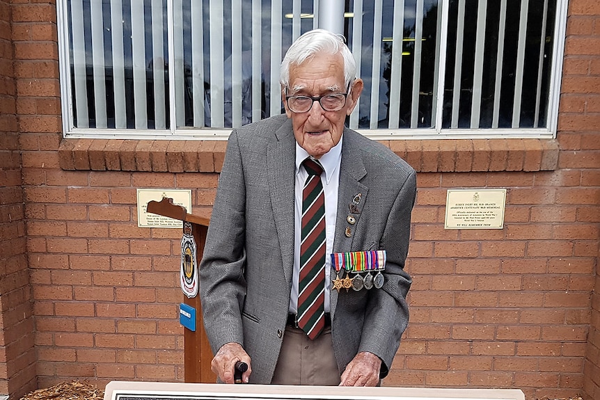 Fred Power stands with a walking stick in front of an Anzac memorial.