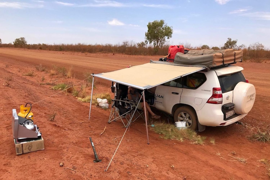 Peter and Sandra Doherty sit in broken down 4WD with awning out, surrounded by red dirt.