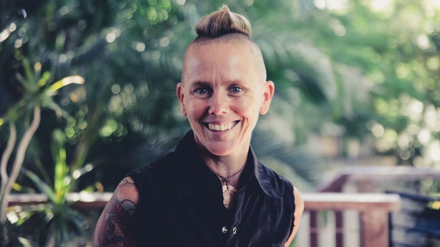 Small-framed person smiling at the camera. They have a mohawk and are wearing a black shirt with cutoff sleeves and tattoos. 