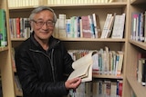 A Chinese man holds a book and smiles with book shelves behind him