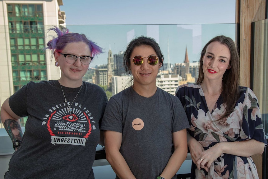 Three people stand on a balcony, with a city skyscape behind them.