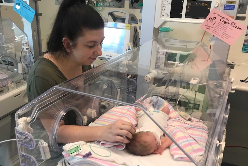 A mother holds a baby girls hand, as the baby lies in a hospital humidicrib.