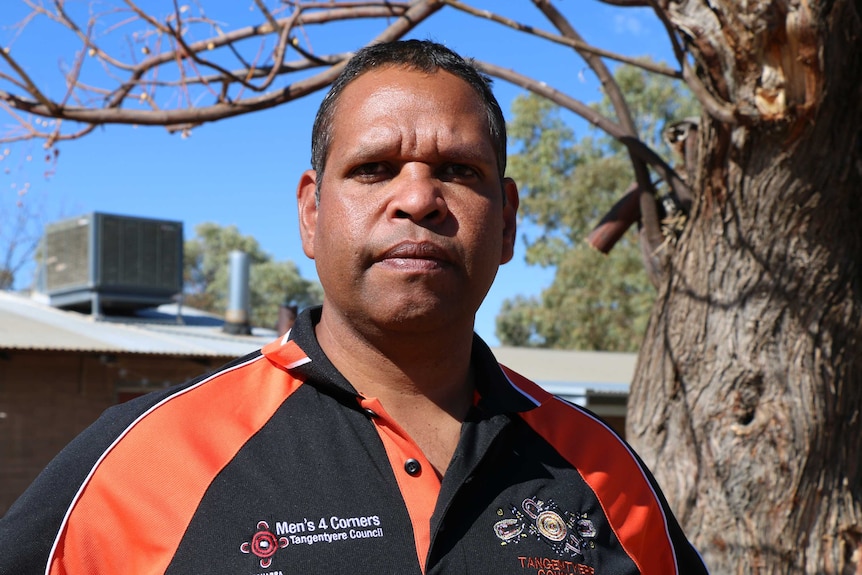 An Indigenous man with short hair, wearing a branded polo shirt and standing near a skeletal tree, looking sombre.