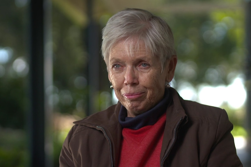 Older woman wearing a red shirt and brown jacket looking at interviewer.