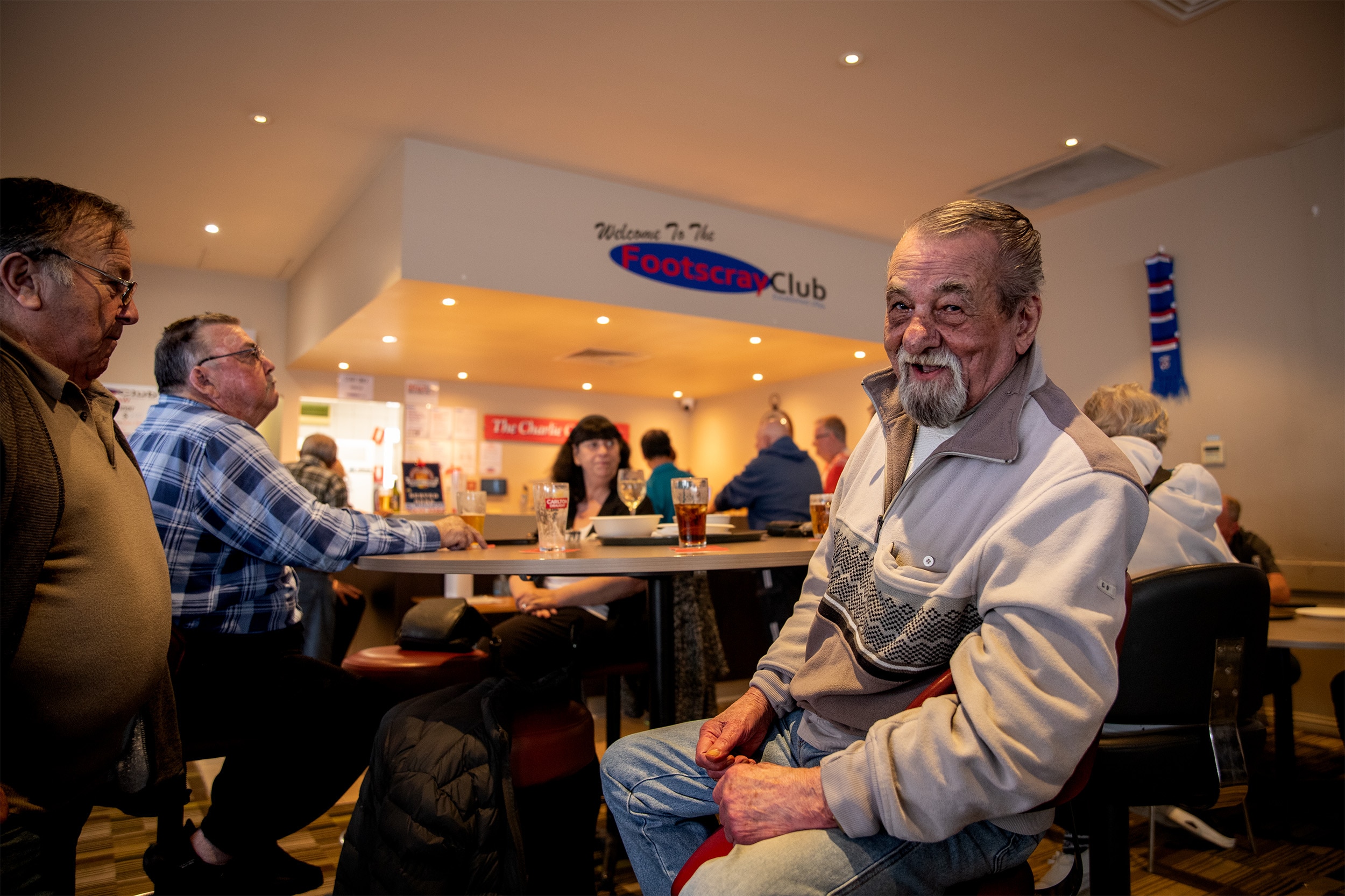 A group of people sit around a table with drinks on it. One man looks at the camera and smiles. He has a grey moustache.