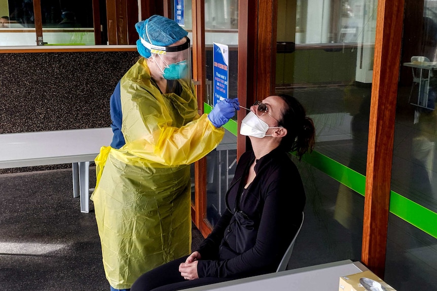 Woman seated wearing black and a white face mask has swab inserted into her nostril by health worker wearing yellow gown and PPE