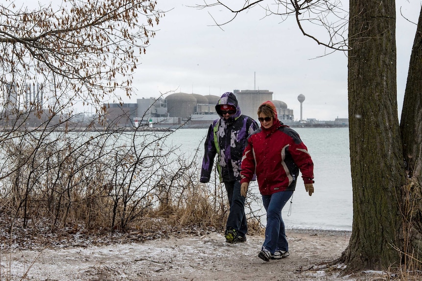 two people walk along snow covered ground in the foreground as a nuclear plant is seen across a body of water behind them