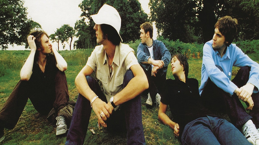 Five members of The Verve sit on the grass in a park with their heads tuned.