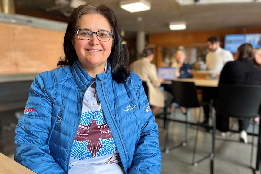 A woman with glasses and shoulder-length hair smiles into the camera, while sitting in front of a table of students.