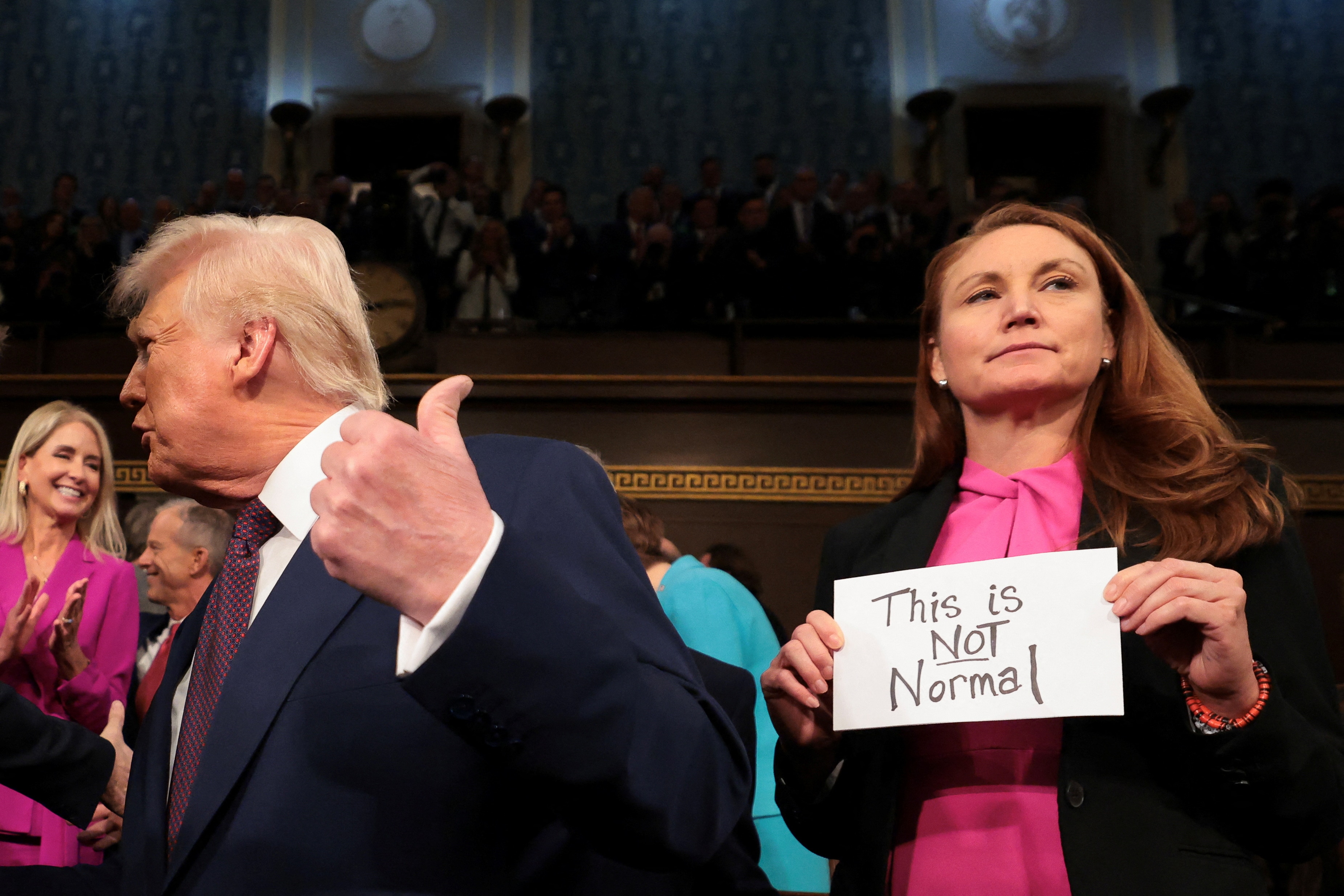 A woman holds a sign reading "THIS IS NOT NORMAL" next to a blonde man in suit 