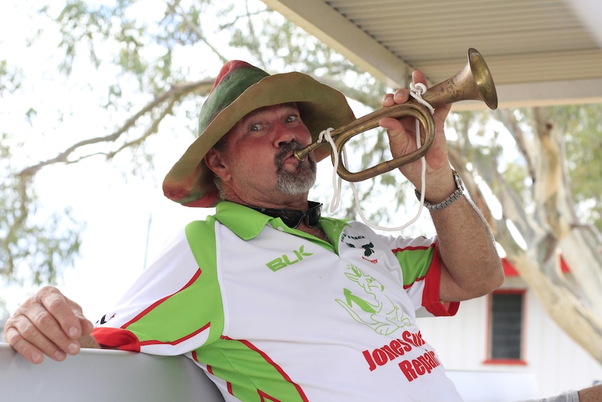 A man in a red and green felt hat holds a bugle in his hand with a grin.