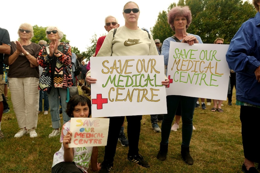 Protesters hold signs in a park urging a GP clinic to stay open