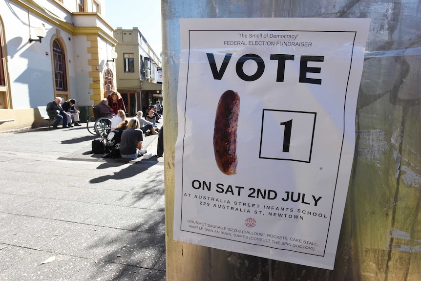 A poster on a pole with the words 'vote 1' next to an image of a sausage. In the background a crowd sits outside in a street.