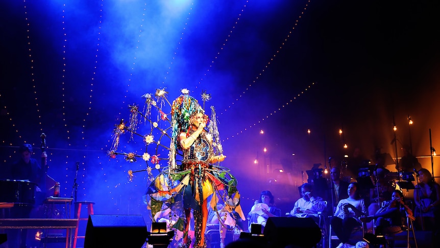 Taylor Mac, a white queer drag performer, sings on a blue-lit stage in an elaborate and colourful costume.
