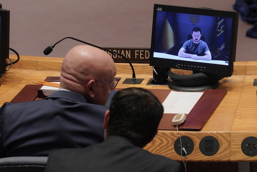Two men chat while watching a monitor on which a third man, dressed in khaki, speaks with arms folded on a desk.
