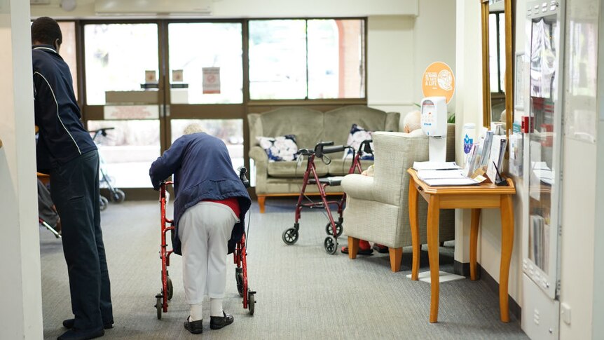 An elderly woman bending over a walker in a nursing home.