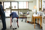 An elderly woman bending over a walker in a nursing home.