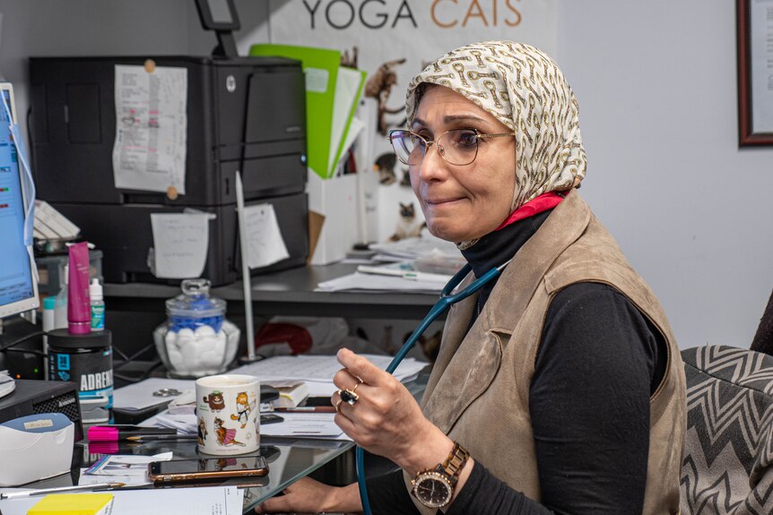 A woman wearing a patterned hibab with a stethoscope around her neck sitting at her desk in her GP rooms.