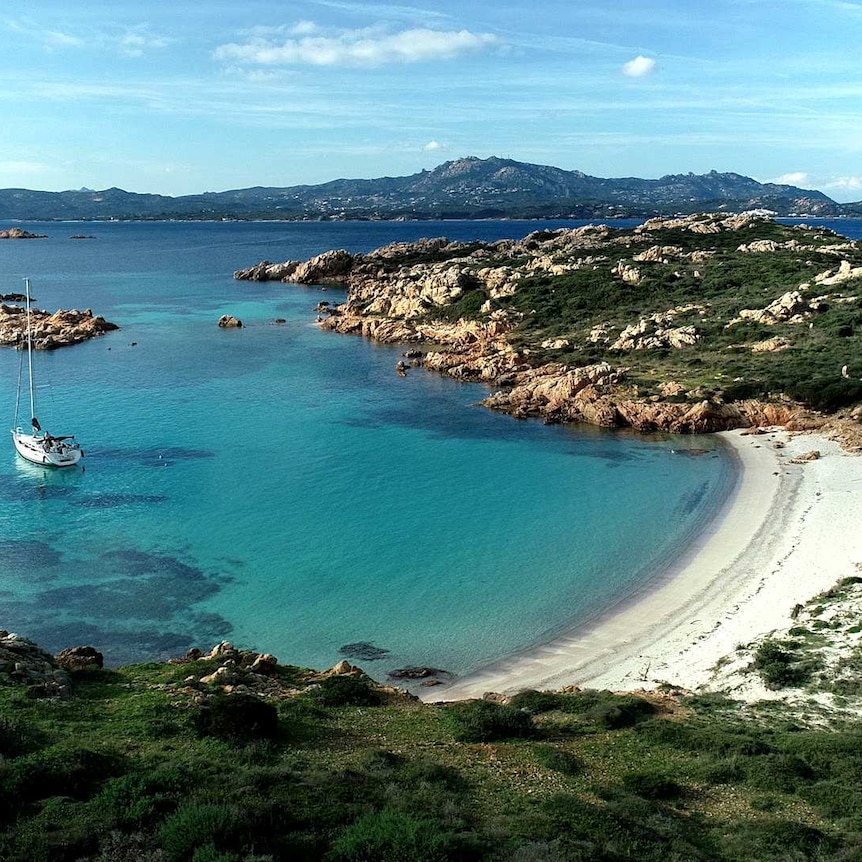 A yacht anchored in a deserted bay filled with bright blue water and bordered with rocky cliffs.