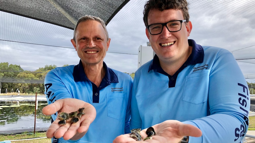 Two smiling men hold up tiny oyster babies called spat on their hands, both wear blue fisheries t-shirts, pond behind.