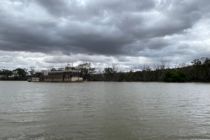 A large paddle boat is in the distance, sitting in murky river water with a gloomy overcast sky.