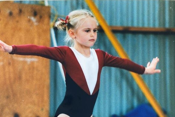 A child with blond hair in pig-tails and wearing a marrone leotard balances, standing with arms outstretched, on a beam.