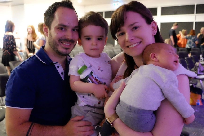 A man and woman, each holding a child, look at the camera and smile.